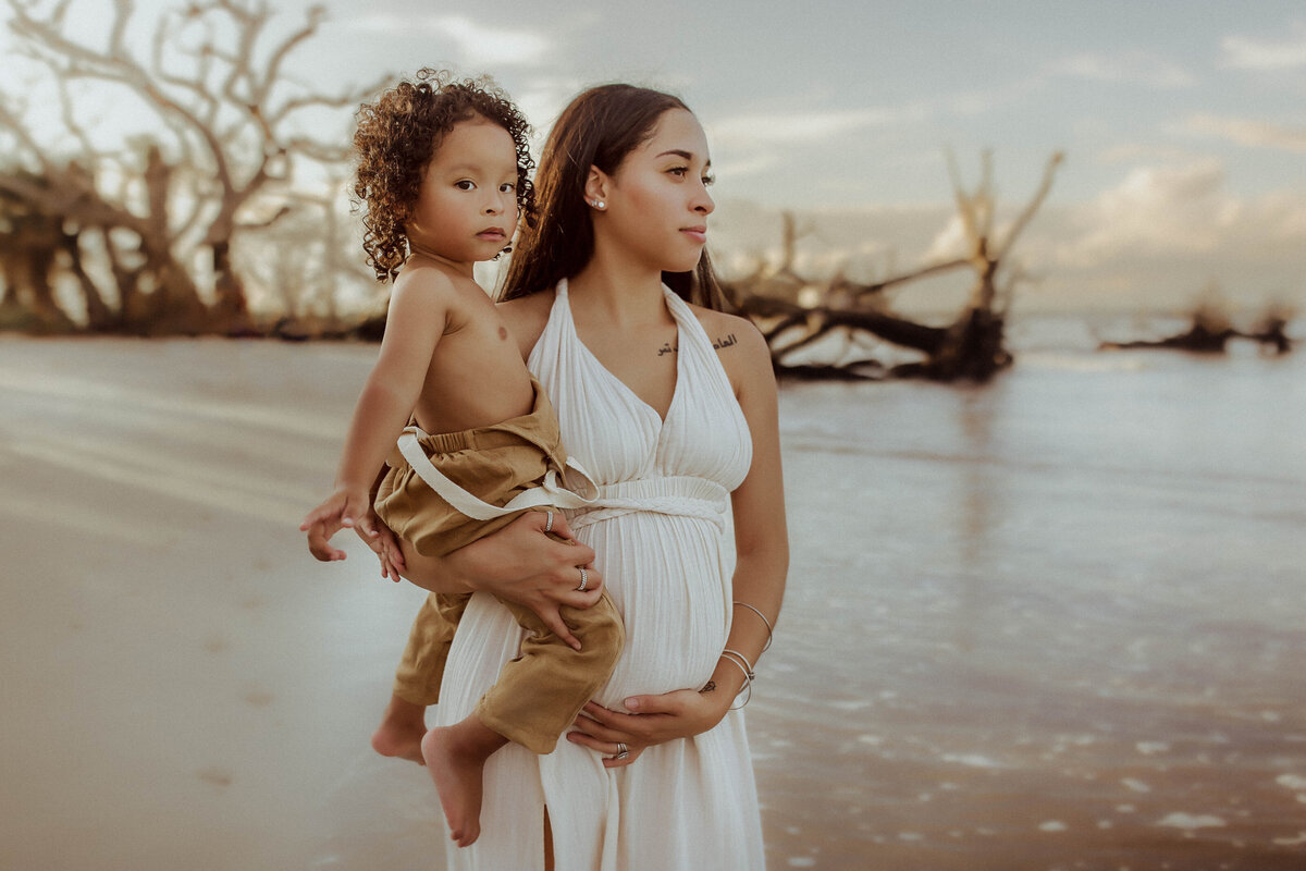 A pregnant woman in a white dress holds a child on a beach with driftwood. They both gaze into the distance.