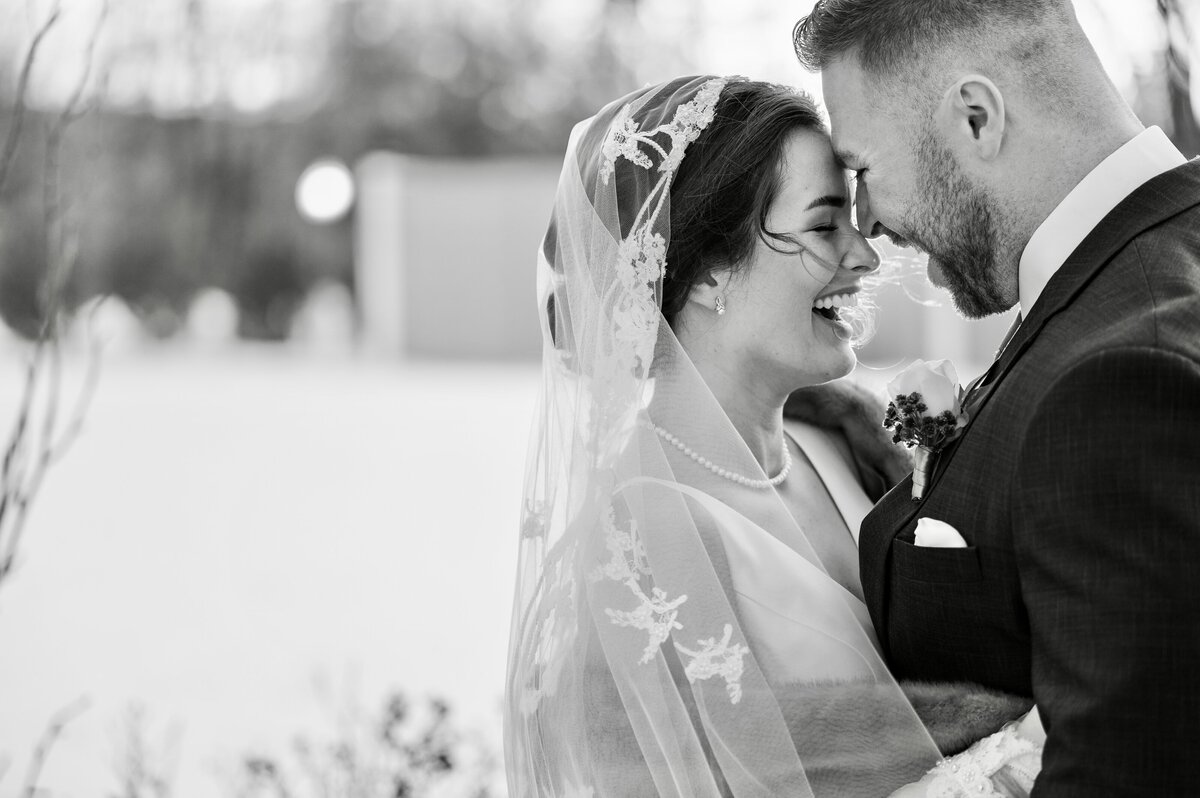 A bride and groom smile as they lean their foreheads together outside the Loch Aerie Mansion in Malvern PA. The bride wears a veil with floral lace, and the groom is in a dark suit, perfectly captured by their winter Philadelphia wedding photographer.