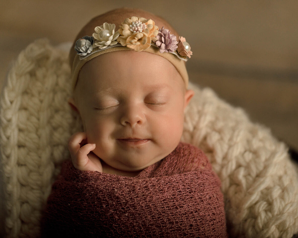 Newborn girl in a dark pink wrap and flowered headband sitting in a wooden bucket.