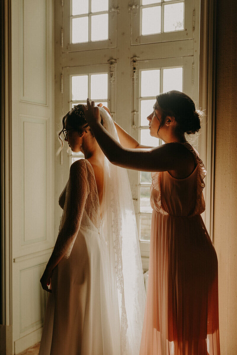 Demoiselle d'honneur accorchant le voile de la mariée devant une encadrure de fenêtre pour une séance photo mariage en Vendée.