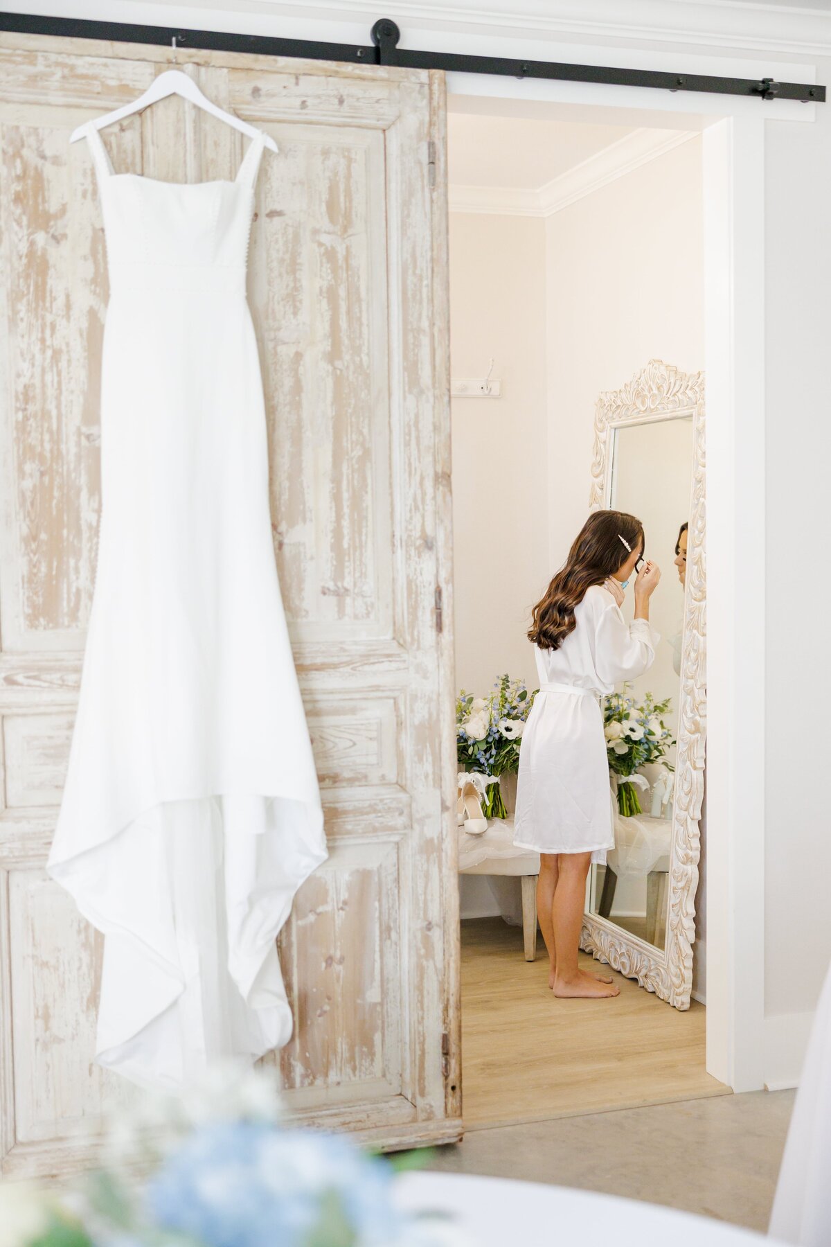 A bride stands in her robe putting on makeup with her wedding dress hanging on the door.