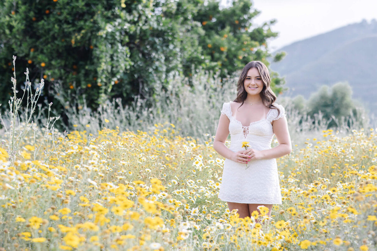 A woman in a white dress stands smiling in a field of yellow and white flowers. She is holding a small bouquet. In the background, there are green trees and a distant mountain under a cloudy sky.