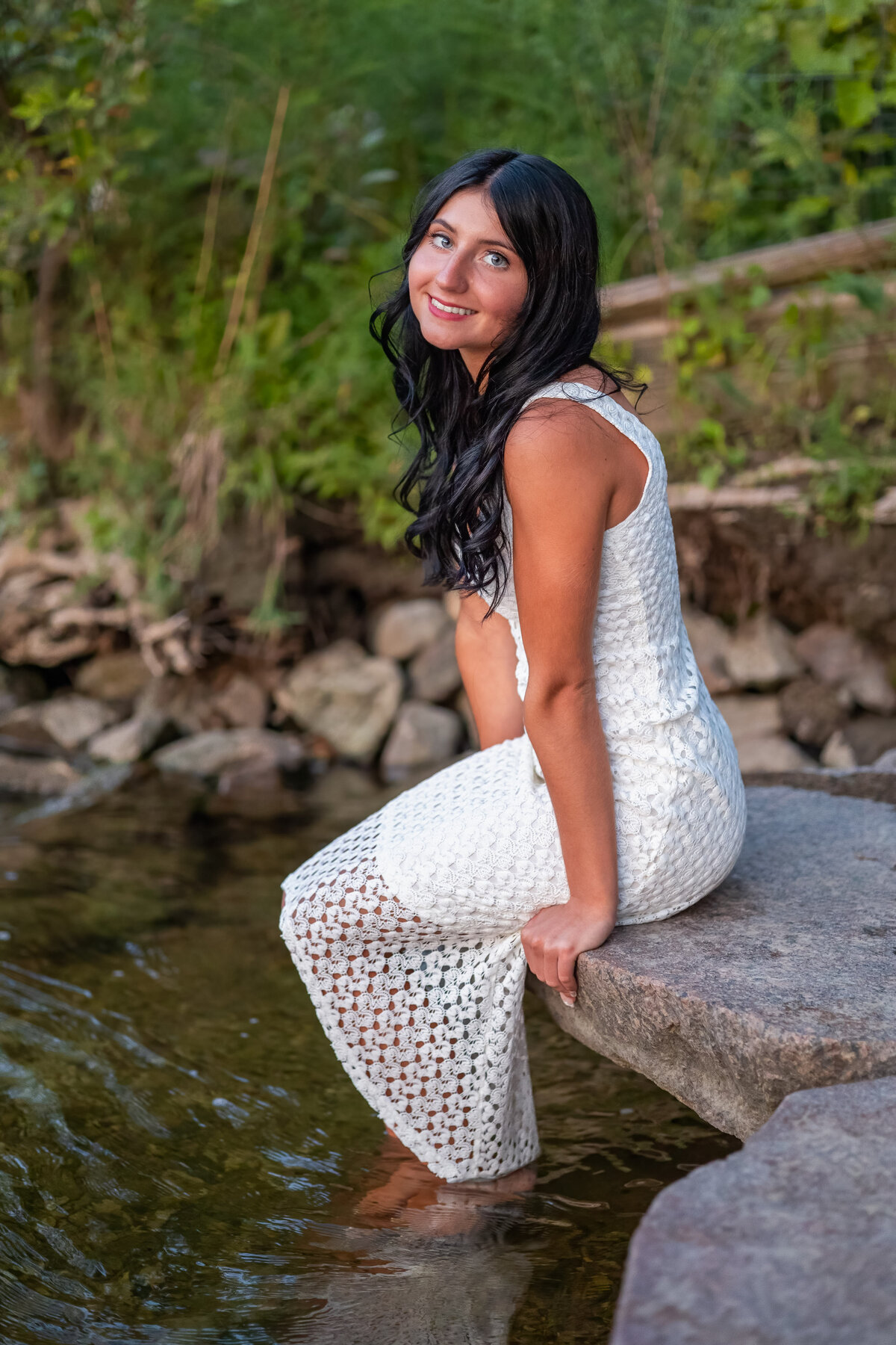 A female high school senior is sitting the edge of a rock dangling her feet into a body of water.  She is looking over her left shoulder looking at the camera and is wearing a long white dress