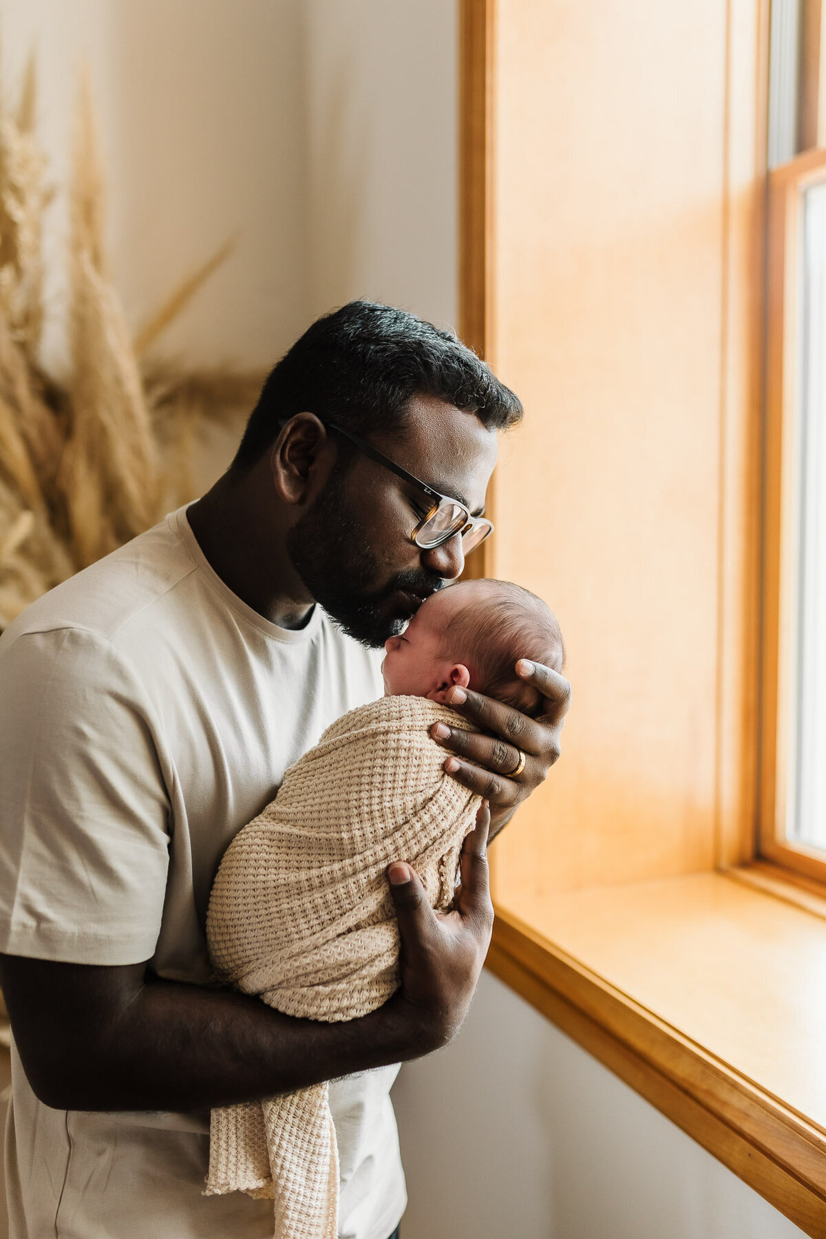 Dad holding newborn baby wrapped in swaddle near a window, kissing his forehead 