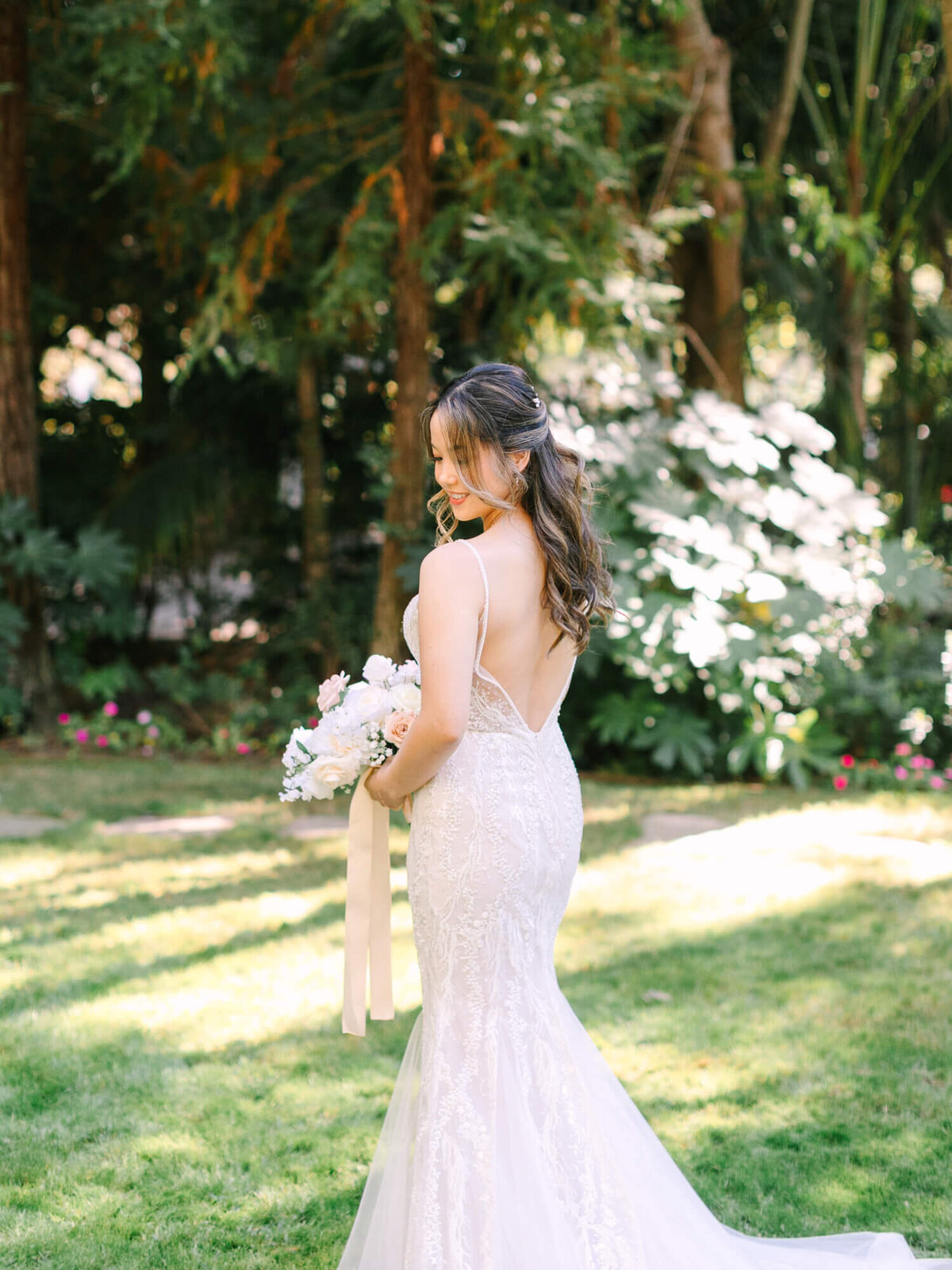 Bride in a lace wedding dress stands in a sunlit garden at Hartley Botanica, holding a bouquet with pastel flowers.