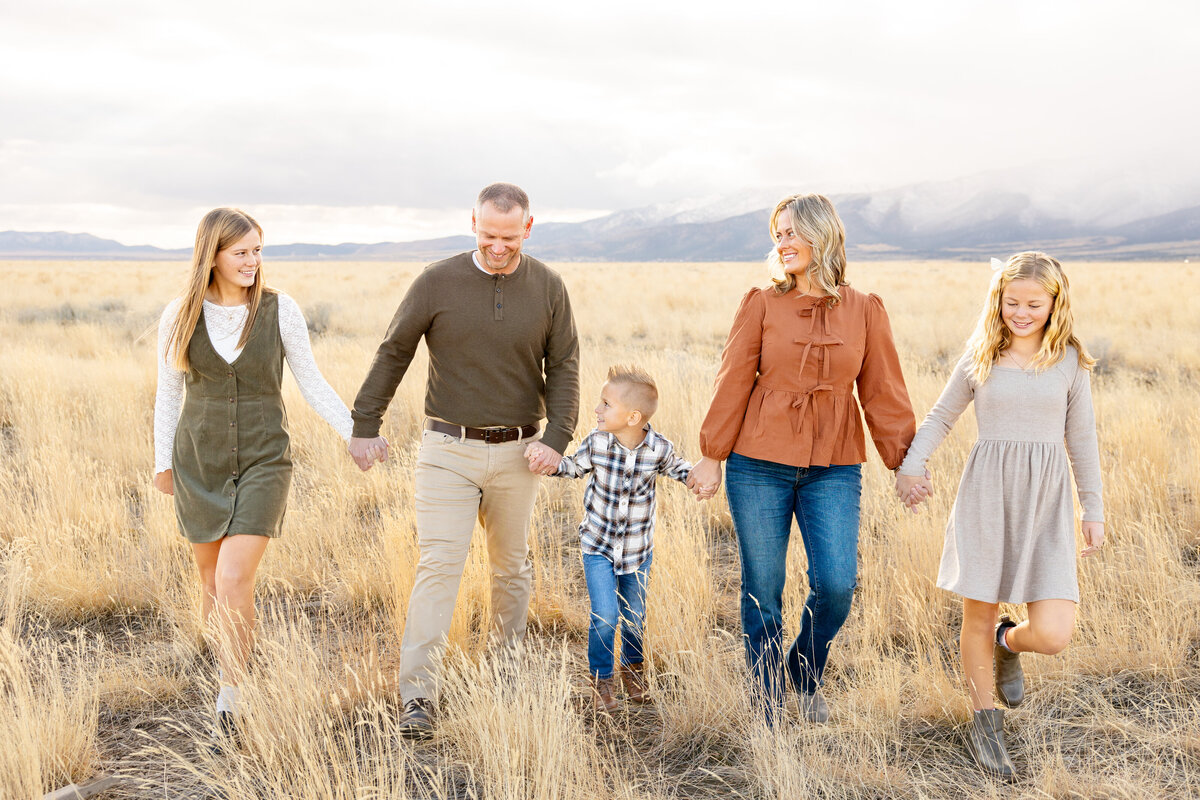 A family of five walks hand-in-hand through a golden field in Eagle Mountain, Utah, with mountains in the background. They are dressed in warm, earthy tones, smiling as they enjoy the outdoor setting.