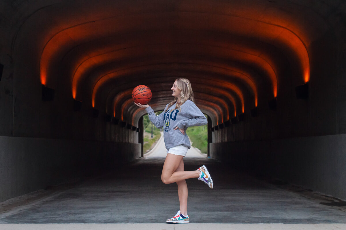A person stands in a dimly lit tunnel, balancing a basketball on their finger. Theyre wearing a gray hoodie and white shorts, with one leg raised. The tunnel has glowing orange lights on the ceiling.