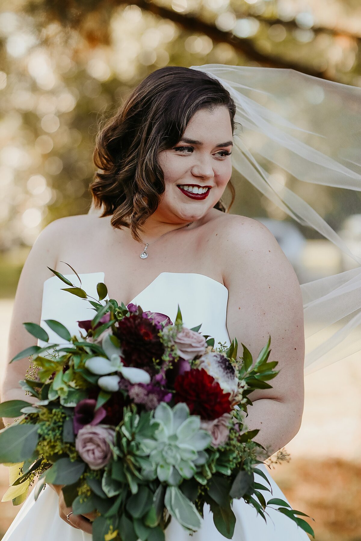Bride wearing a sheer veil, dark lipstick and a strapless wedding dress with a v neckline holds a bouquet of succulents, burgundy dahlia, lilac roses and eucalyptus.
