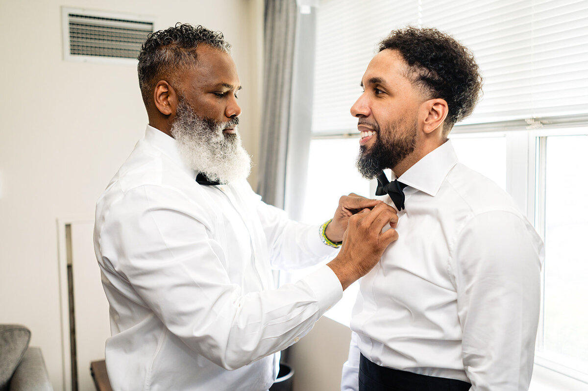 A heartwarming moment between two men in a bright room, with an older man adjusting a younger man's bow tie, both dressed in formal white shirts