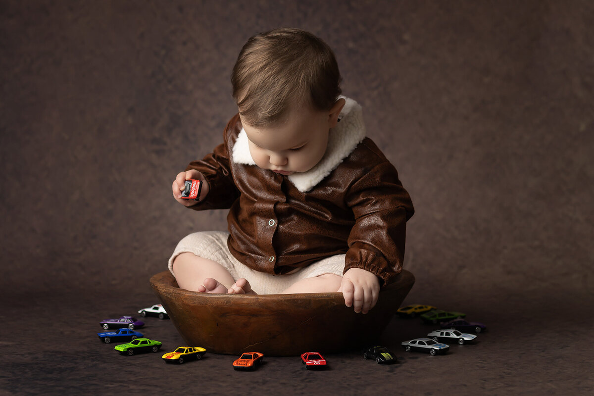 Toddler in a brown aviator jacket sitting in a wooden bowl, looking down while holding a red toy car, surrounded by colorful toy vehicles, styled by a Dallas milestone photographer.