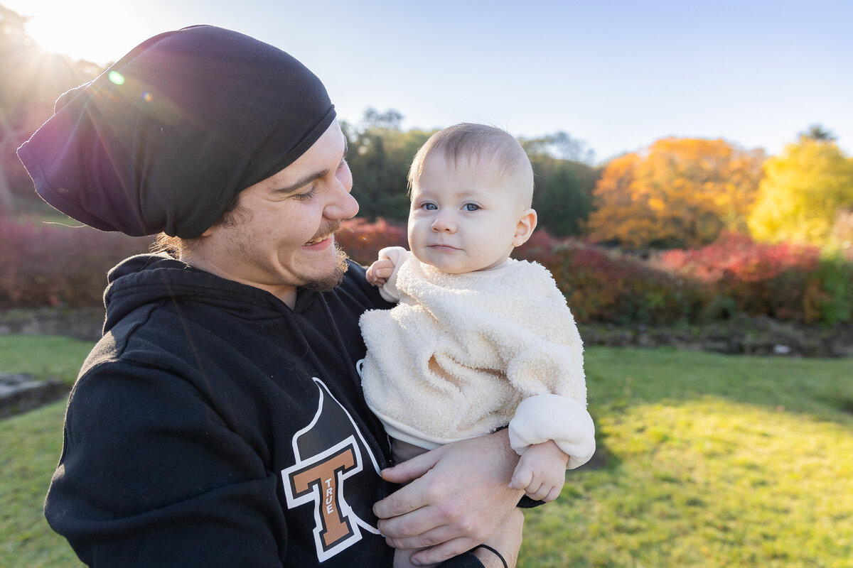 A young man in a beanie holds a baby wearing a white fluffy outfit, both smiling in a park with autumn leaves in the background.