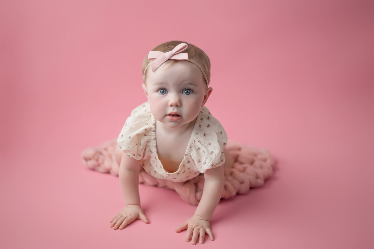 A baby wearing a light dress and pink headband is crawling on a pink blanket against a pink background