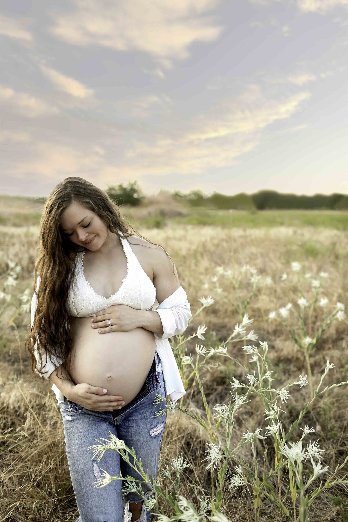 A peaceful maternity photo of the expectant mom standing in a field of white flowers, gently cradling her belly with a soft smile, captured by Chunky Monkey Photography.
