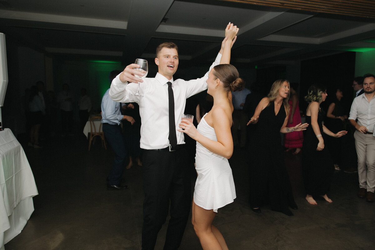 Bride in short, strapless reception dress holds her groom's hand as they face each other dancing and holding their hands high in the air. They both hold whine glasses in their other hands.  The groom is singing along to the music. He is in a white dress shirt, black slacks and a thin long black tie. People dance behind them. 