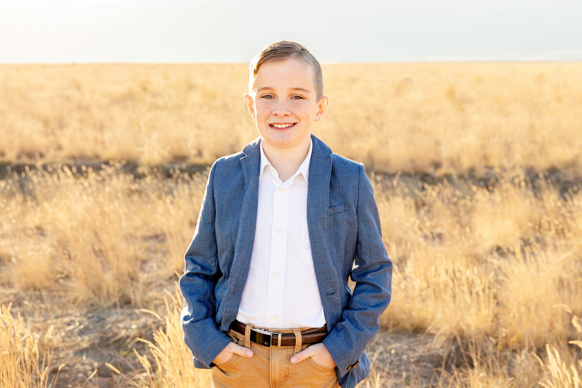 A well-dressed young boy in a blue blazer and white button-up shirt poses in a golden field in Eagle Mountain, Utah, with a bright smile.