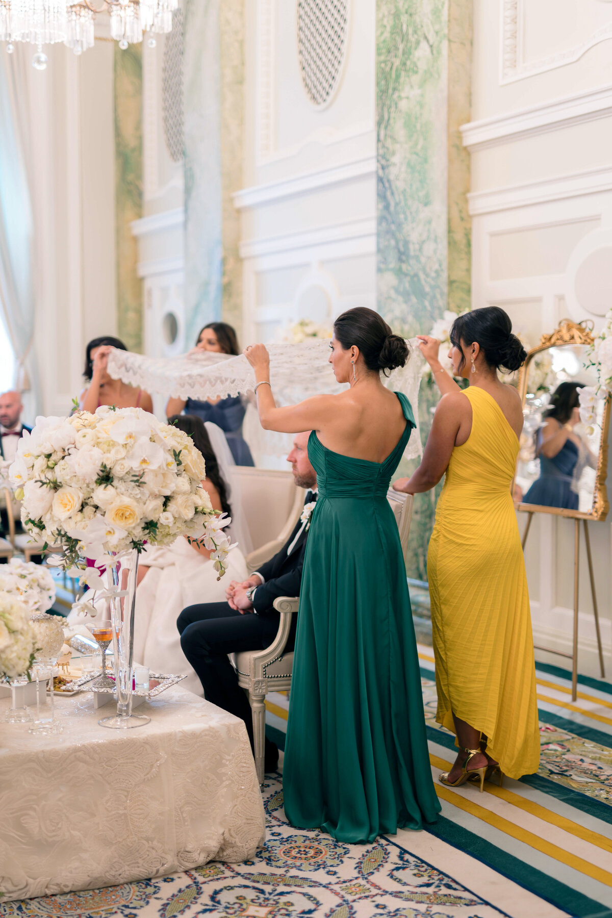 A wedding ceremony scene with a bride and groom sitting beneath a lace canopy held by two women in elegant dresses, one in green and the other in yellow. The room is adorned with floral arrangements and ornate decor.