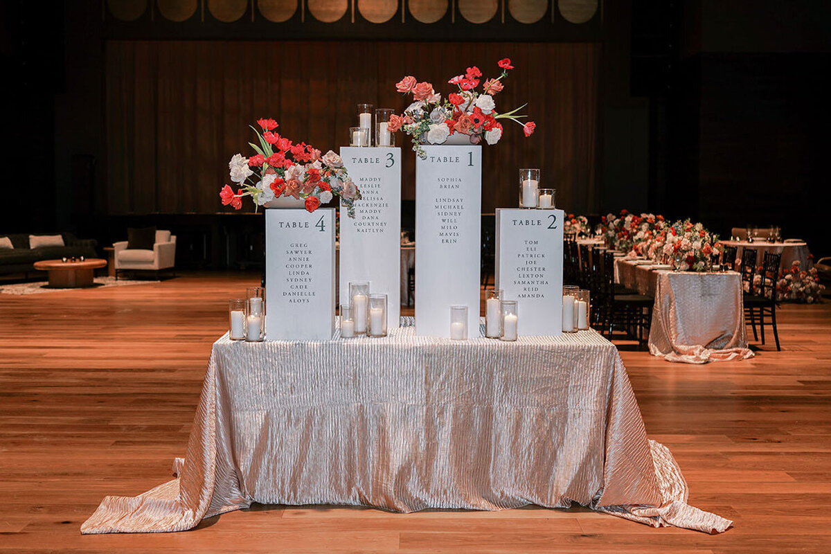 Modern wedding seating chart display with tall white pillars adorned with red, pink, and blush floral arrangements, surrounded by votive candles on a blush textured tablecloth.