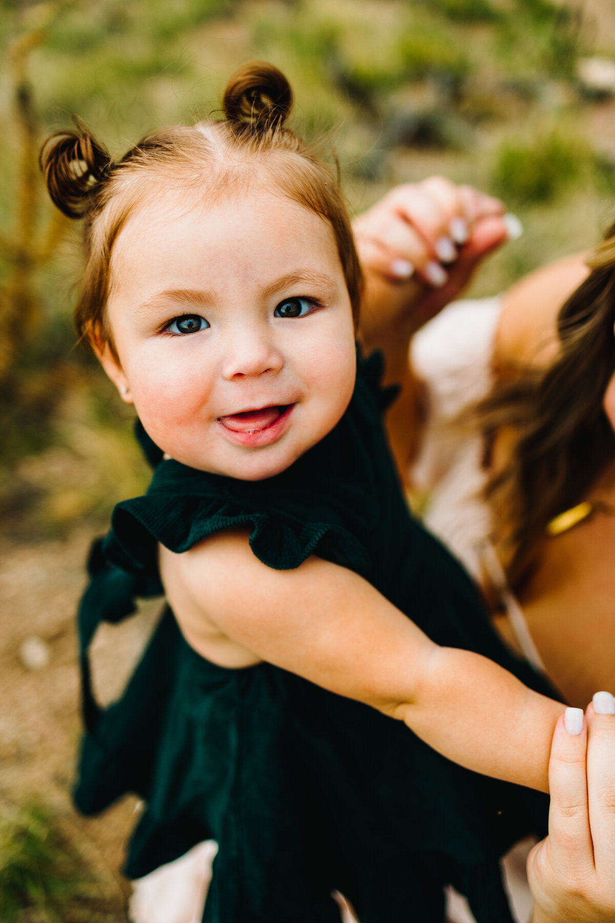 Photo of a baby watching the camera, she has a dark green dress and two tails in her hair. She is probing and is holding a woman's hand