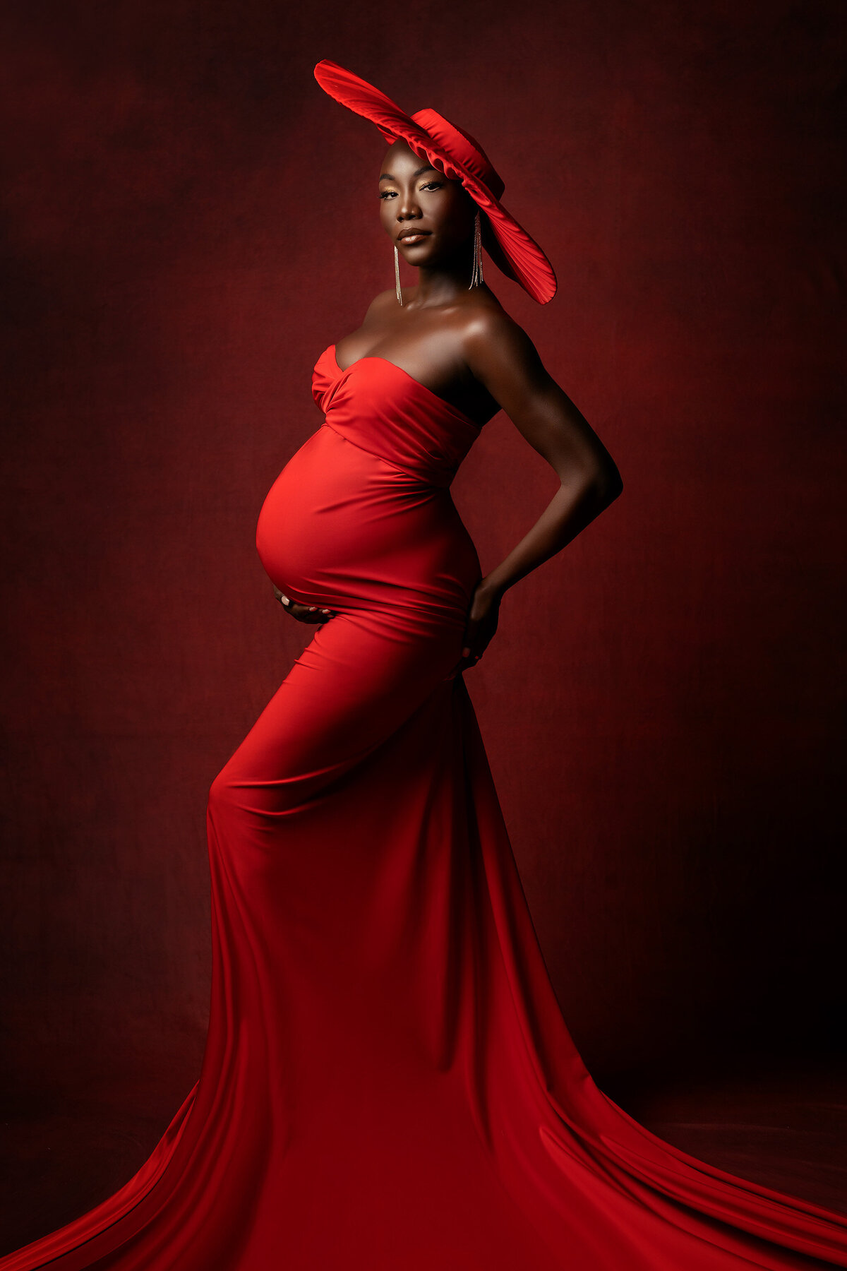 A striking woman draped in a flowing red gown and matching hat poses with elegance against a deep red backdrop, her confident gaze and radiant style embodying the luxurious beauty of fine art maternity photography.