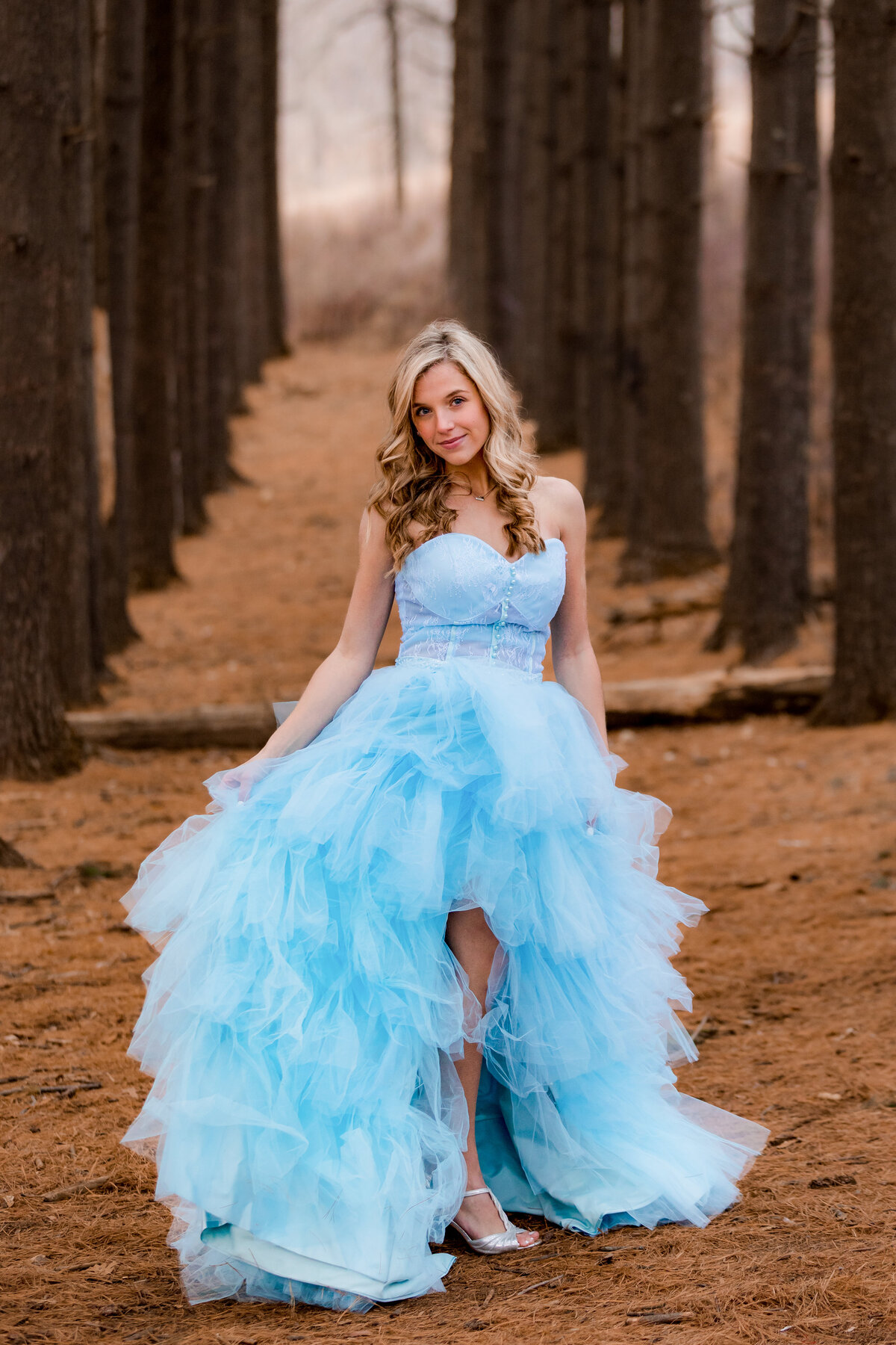 A blonde senior swirls her blue couture gown side to side while standing in rows of trees.