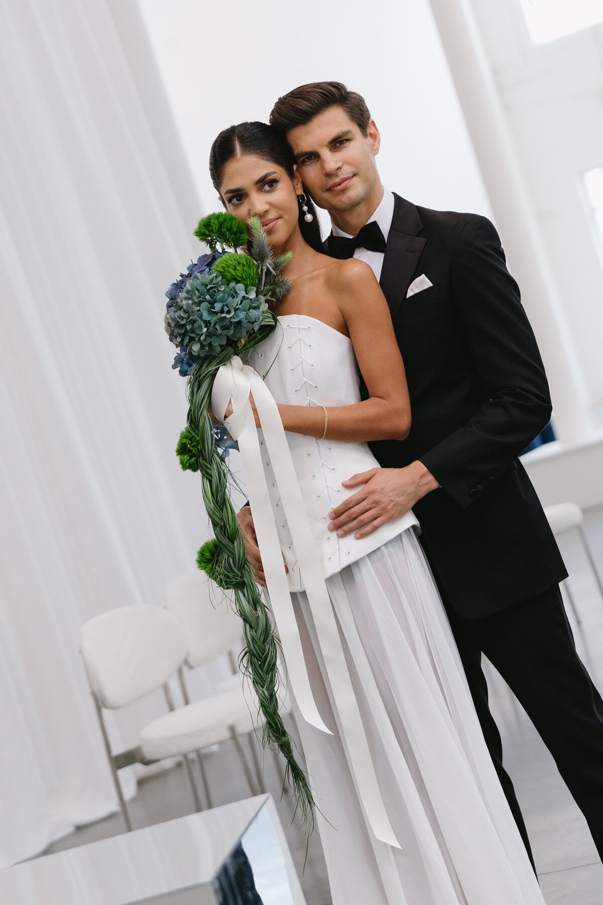 Bride and groom embracing during a modern editorial wedding shoot at Sound River Studios, featuring a sculptural bouquet and clean white backdrop.