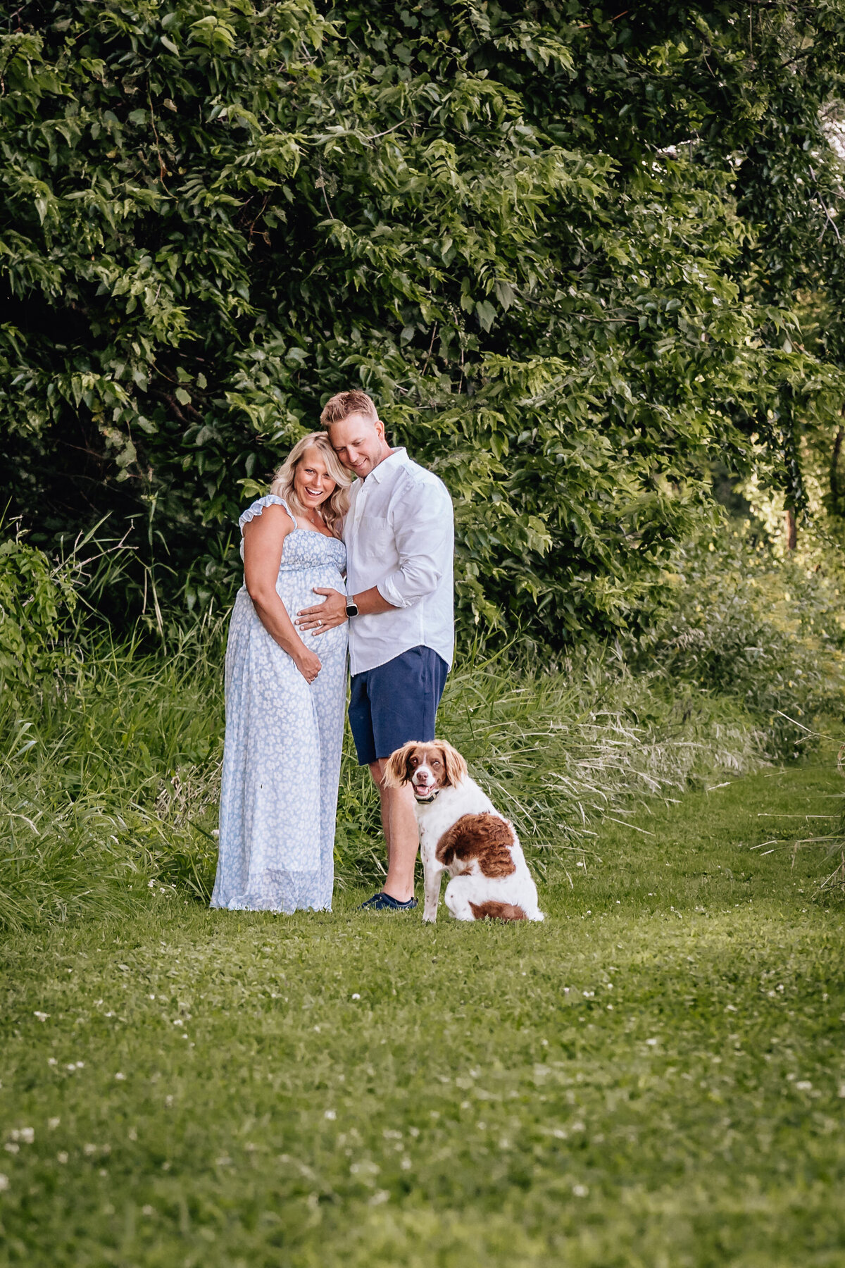 A pregnant couple poses with a dog in a grassy area near trees, the couple embracing while the dog sits happily in front of them.