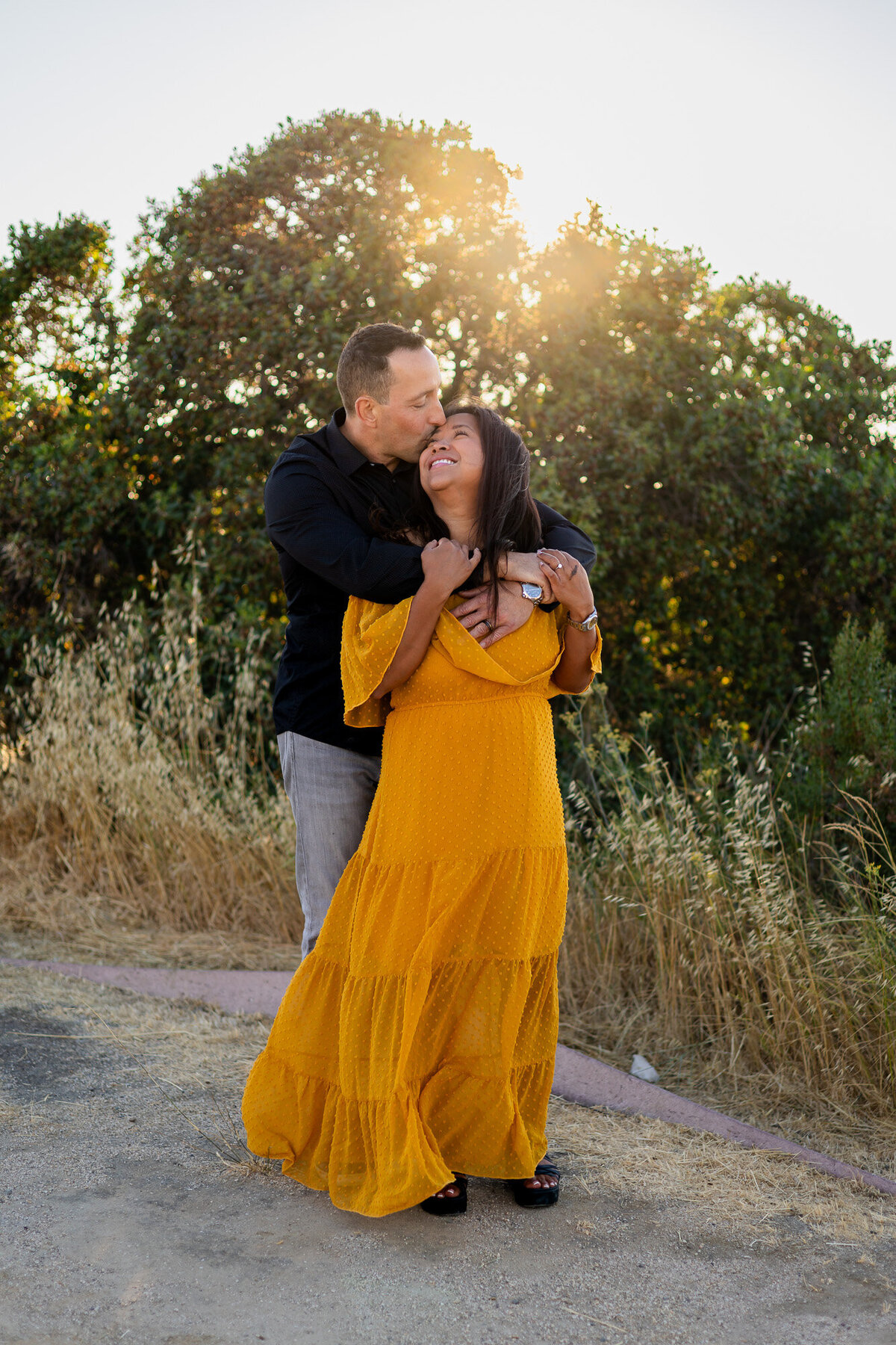 Man embracing his partner with a kiss on her temple as she smiles in front of sunlit trees – Bay Area Couples Photography Portfolio - Ellobelle Photography