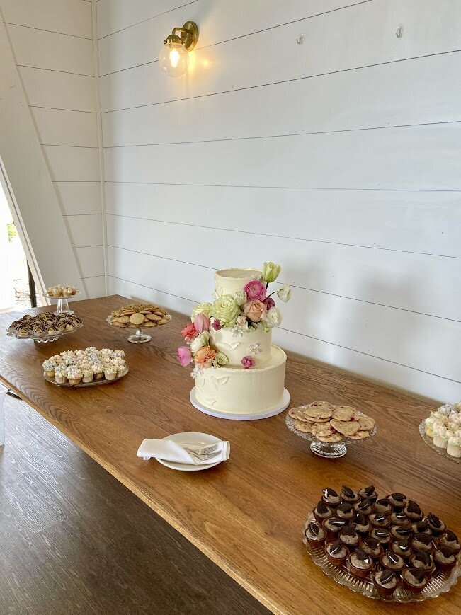A three tier cake with fresh spring florals sits on a large wooden farmhouse table, surrounded by stands filled with mini pastries, cookies, and cupcakes.