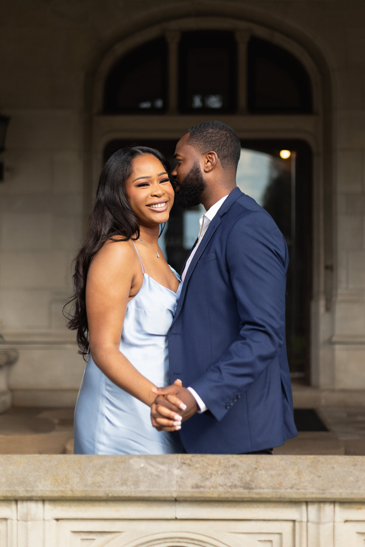 Engagement Photos Rhode Island | Kelsey Sheehan Photography Timeless Rhode Island Weddings | A couple embracing warmly; the woman in a light blue dress smiles joyfully while holding hands with the man in a dark suit standing on a stone walkway.