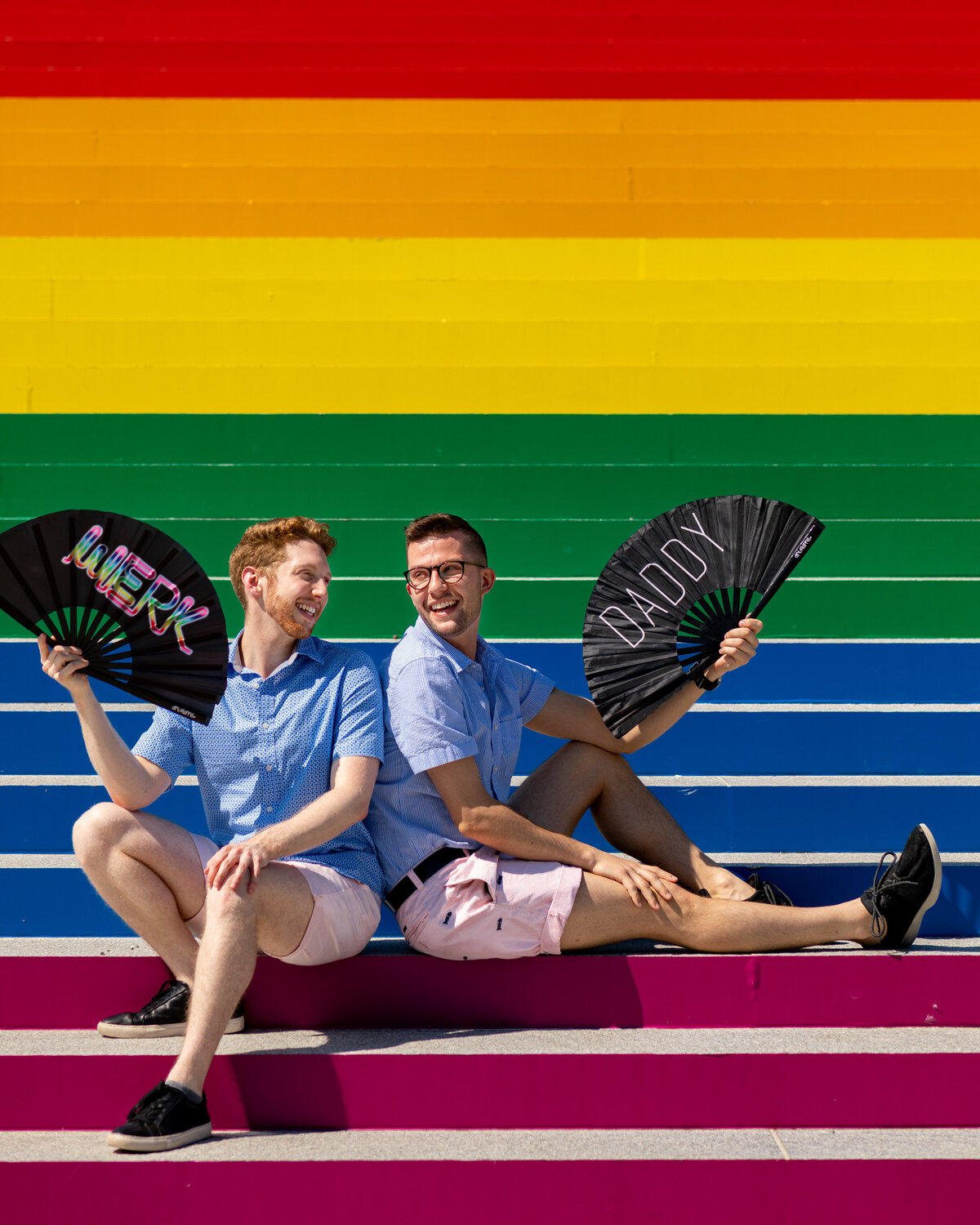 A couple sitting on a rainbow staircase holding decorative fans.