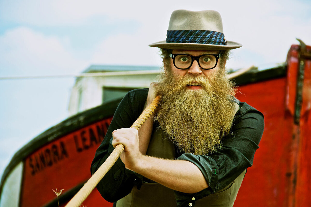 Musician portrait Ben Caplan wearing hat glasses and long beard holding tow rope of red tug boat behind him