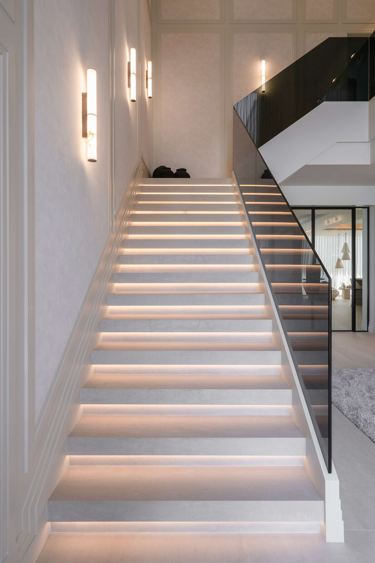 A modern staircase with a minimalist design featuring wide, white steps illuminated by soft, integrated lighting. The stairs are bordered by a sleek, black glass railing on the right side. The walls are adorned with vertical, cylindrical wall sconces that emit a warm glow. At the top of the staircase, black decorative vases are visible, adding a touch of contrast to the light-colored interior.