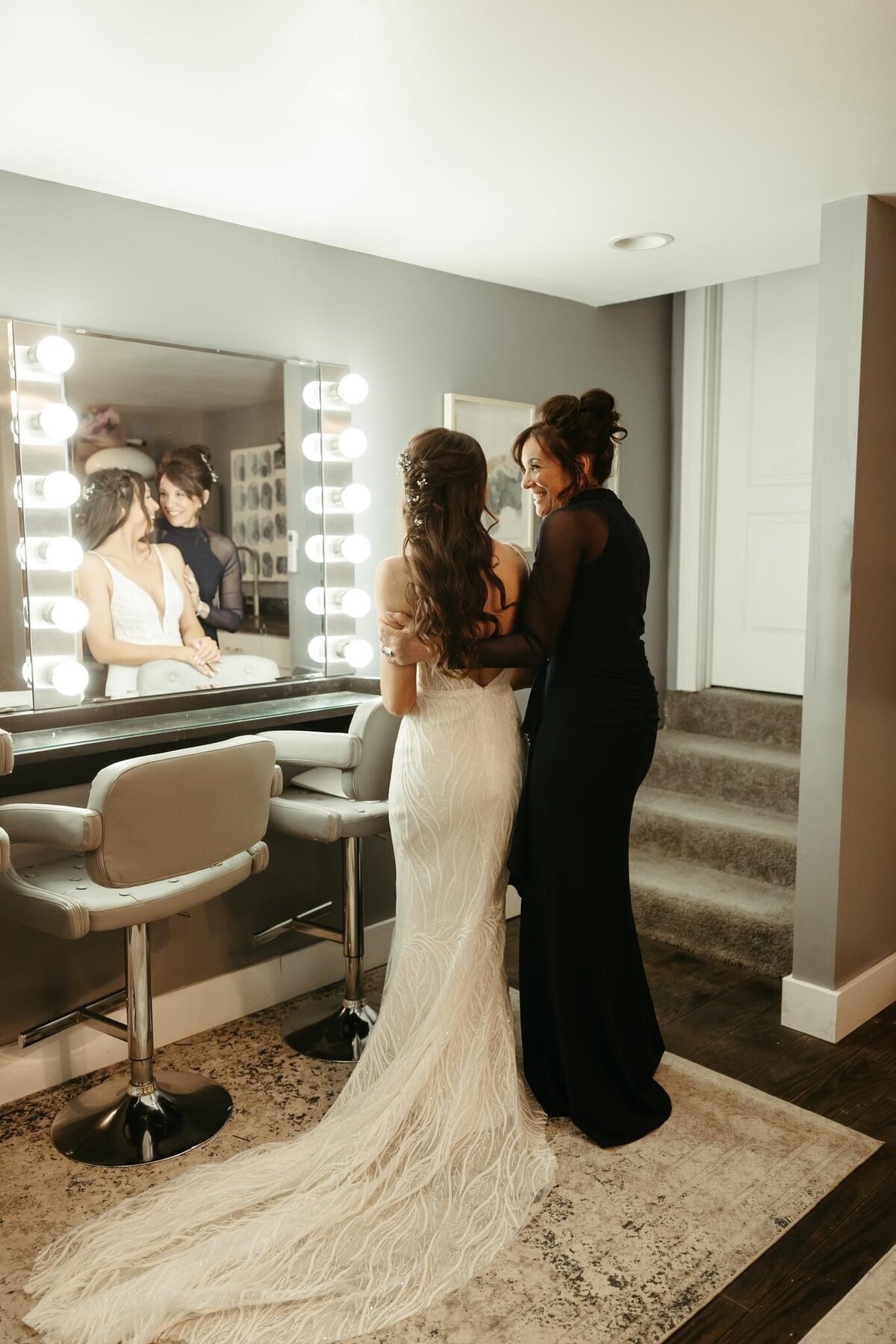 mother of the bride dressed in a navy gown standing with the bride in the Willowbrook wedding venue preparation suite by the lighted make-up mirrors