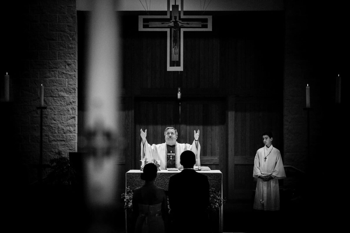 A solemn black and white photo of a priest conducting a wedding ceremony, with a couple in the foreground.