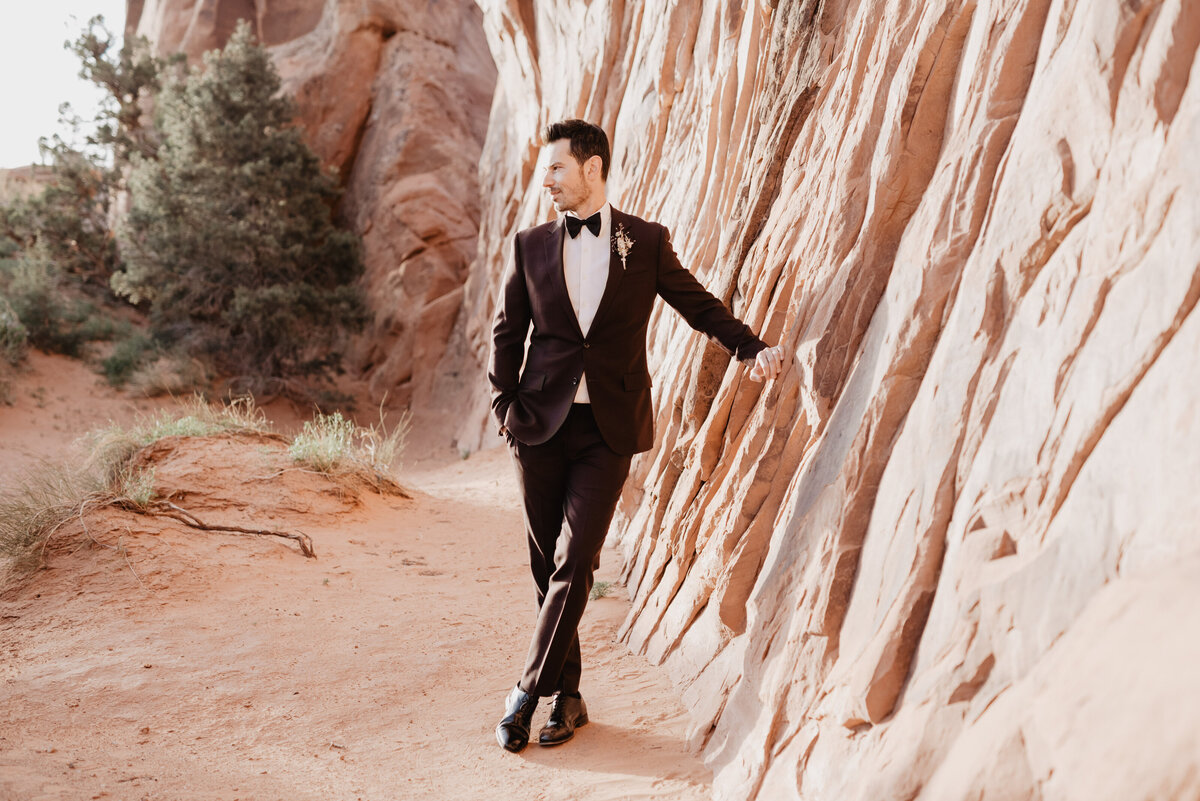 Utah elopement photographer captures groom leaning against rock wearing tuxedo