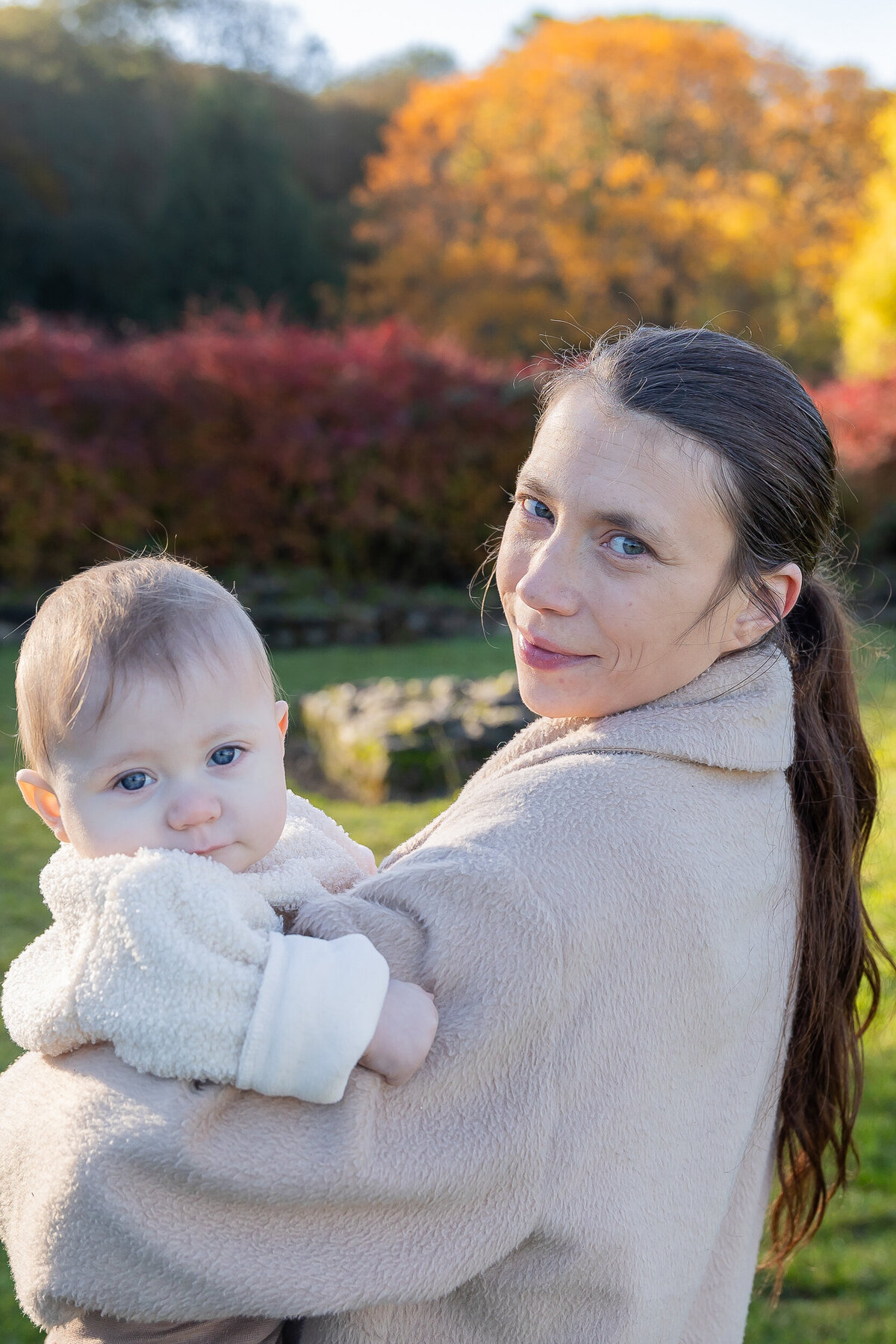 A woman holding a baby, both looking at the camera, with autumnal trees in the background.