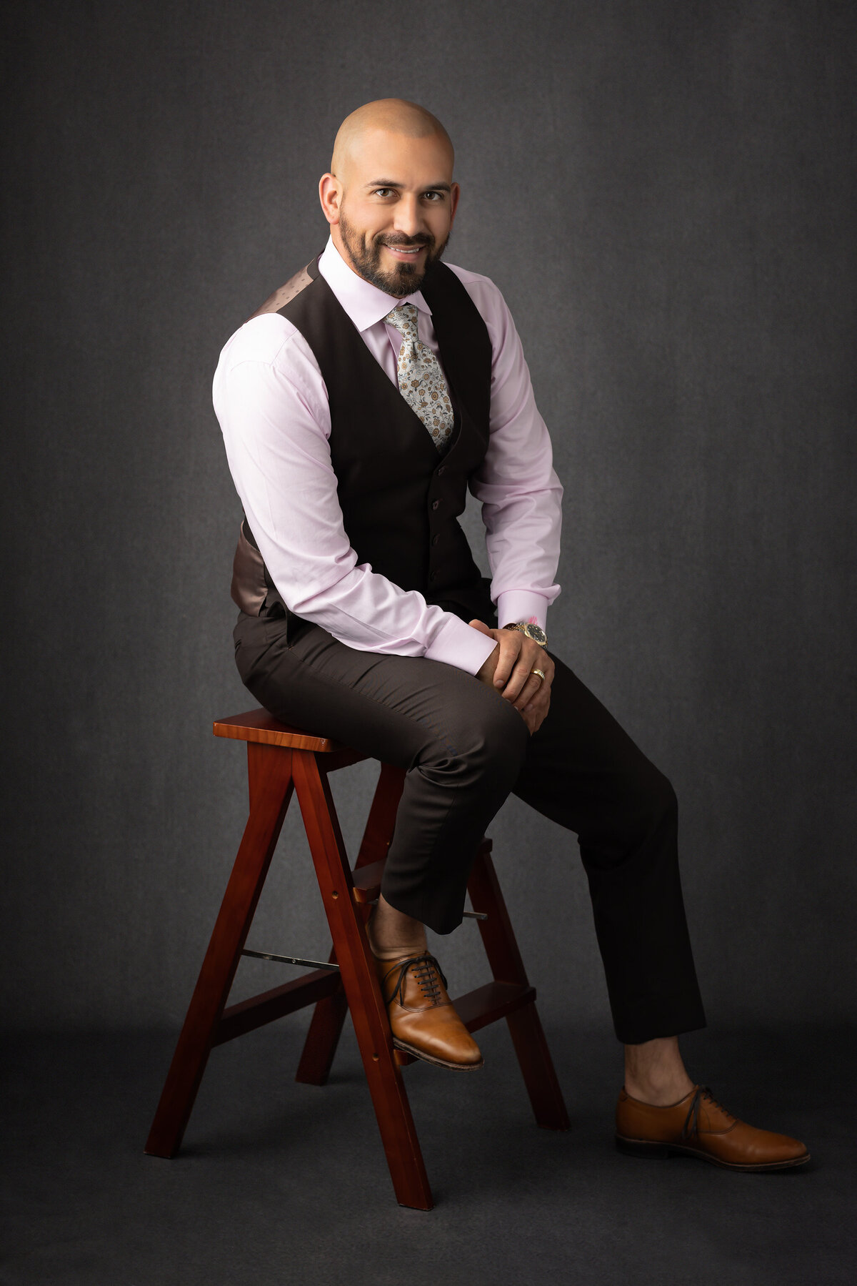 Dallas headshot photographer captures a man in a brown suit, sitting on a wooden stool with a confident smile.