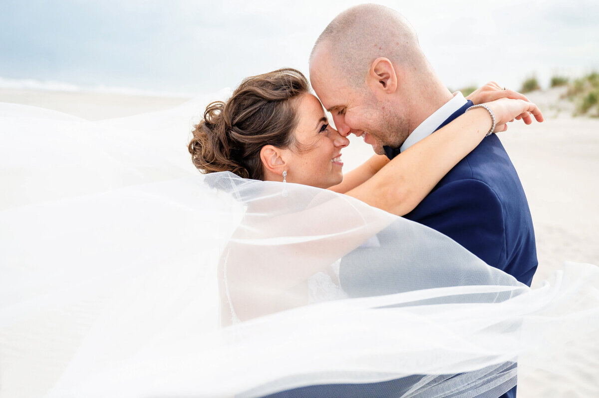 bride and groom, hugging each other, nose to nose with the brides veil, blowing in the wind on the beach before their wedding reception at the yacht club of Stone Harbor