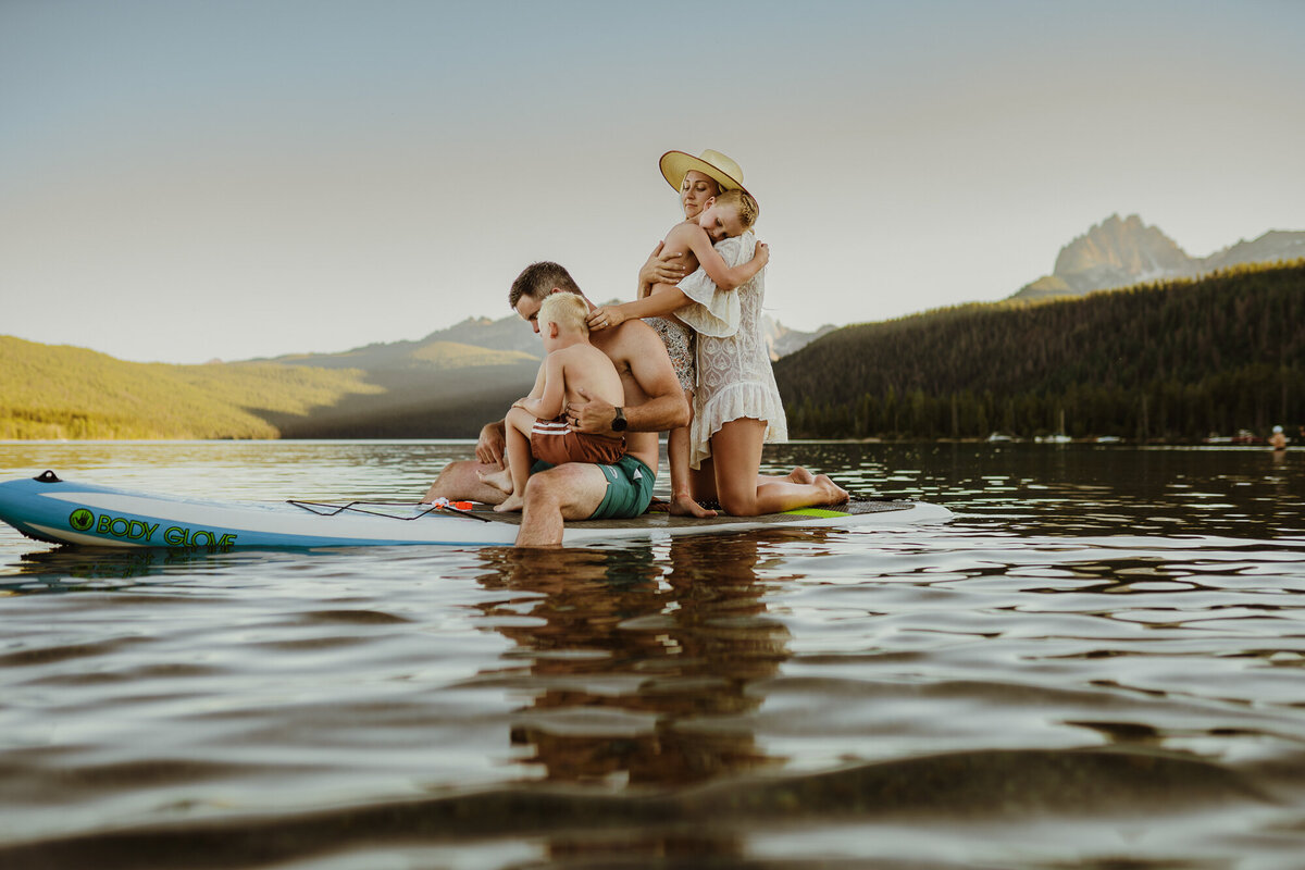 A family of four sits on a paddle board hugging each other  in the lake