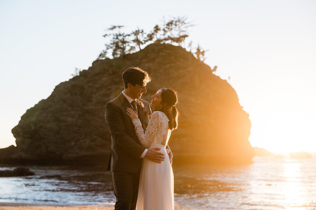 A bride and groom stand on the beach during sunset and laugh.