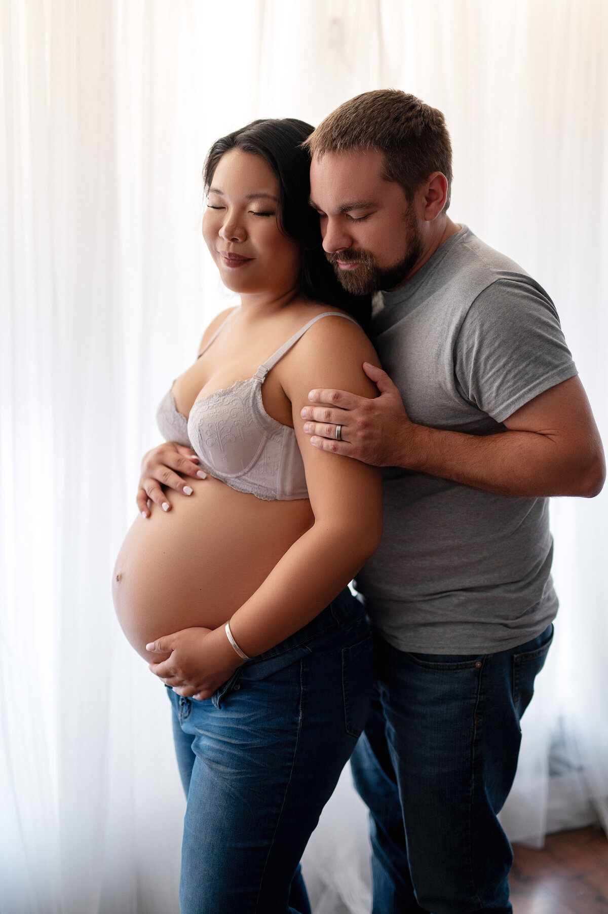 Pregnant woman in a lavender bra and jeans standing in soft window light, with her partner embracing her from behind during a cozy, love-filled maternity session in Waukesha, Wisconsin.