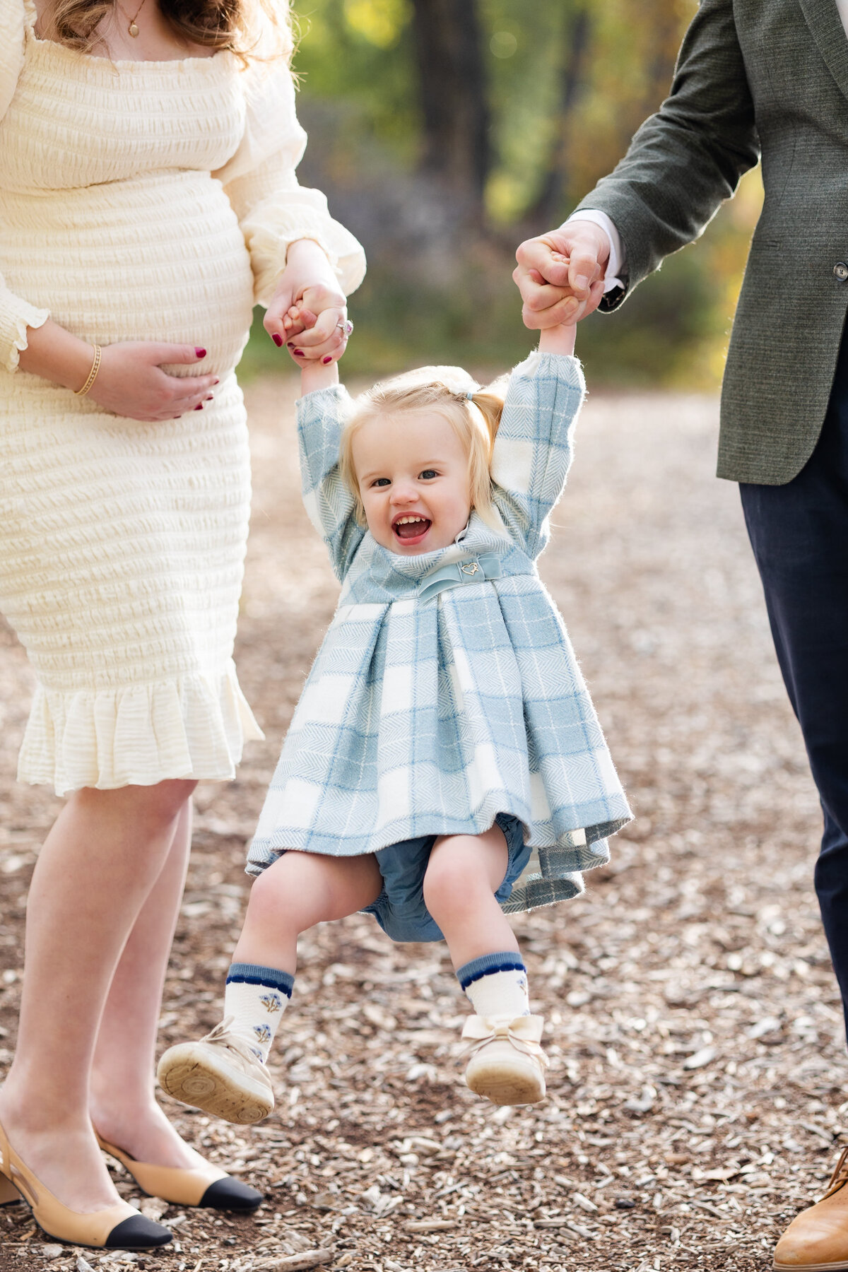 Close-up of toddler girl in a blue and white plaid dress swinging from her parents' hands and laughing.