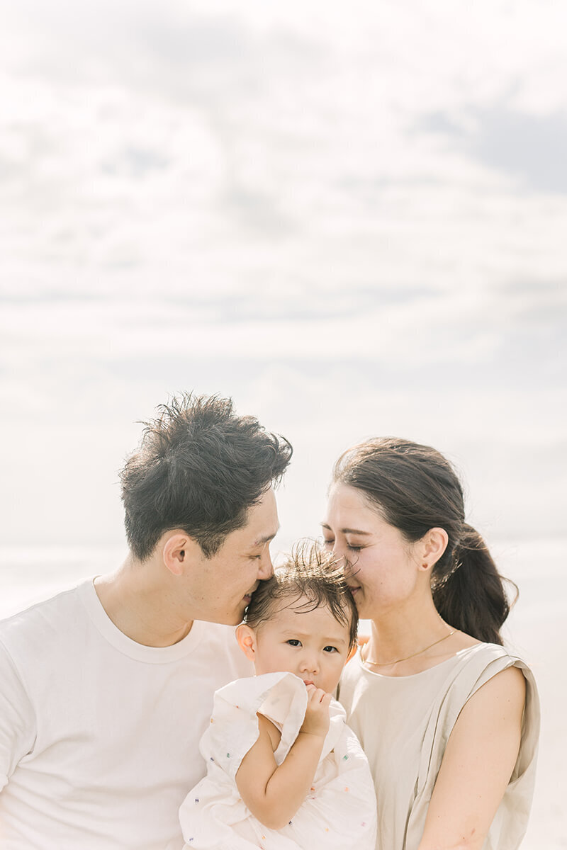 海をバックに、女の子の赤ちゃんにママとパパがキスをしている、美しい家族写真。藤沢市の湘南の海辺でYukika Ishikawa Photographyが撮影。