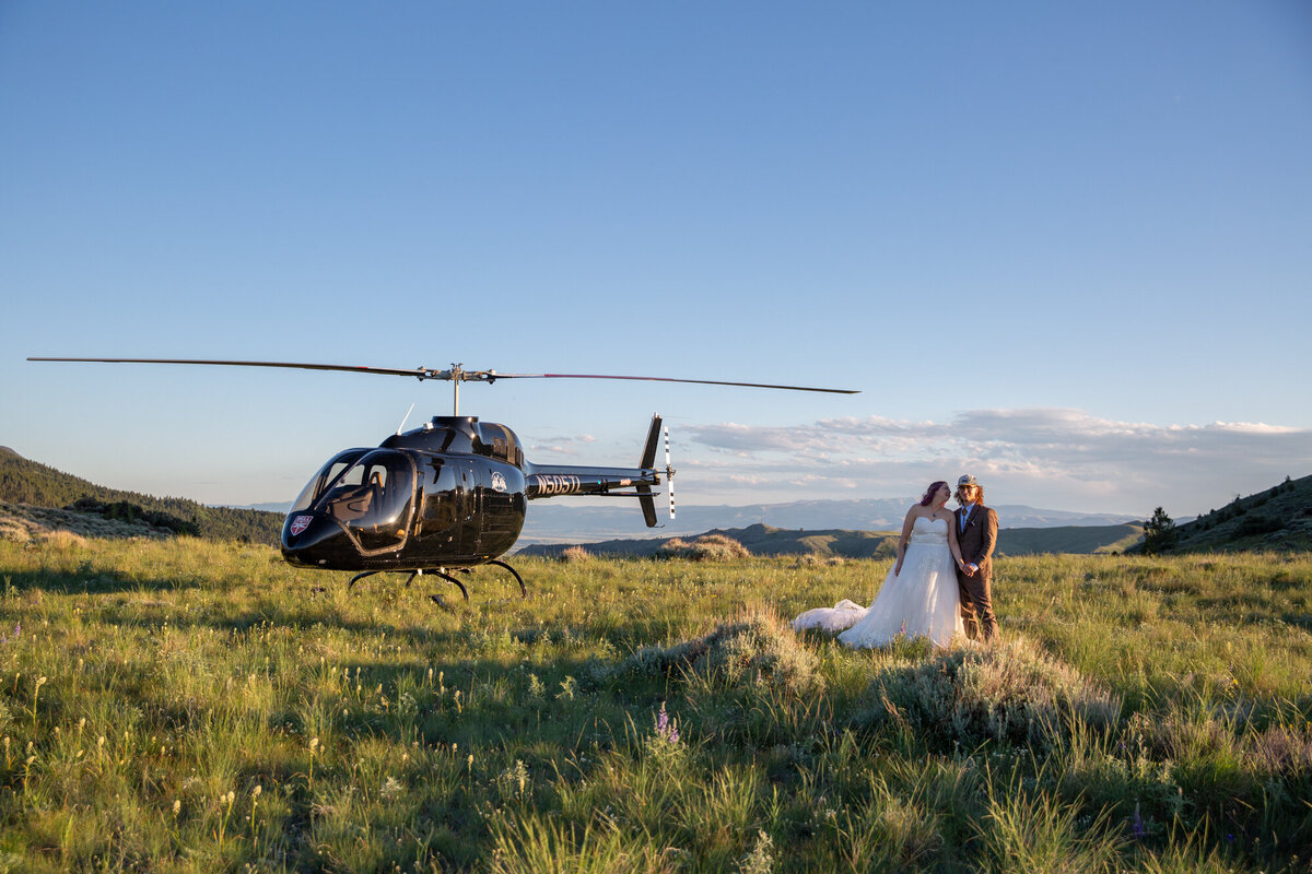 A bride and groom stand in a grassy field next to a parked helicopter on their elopement day.