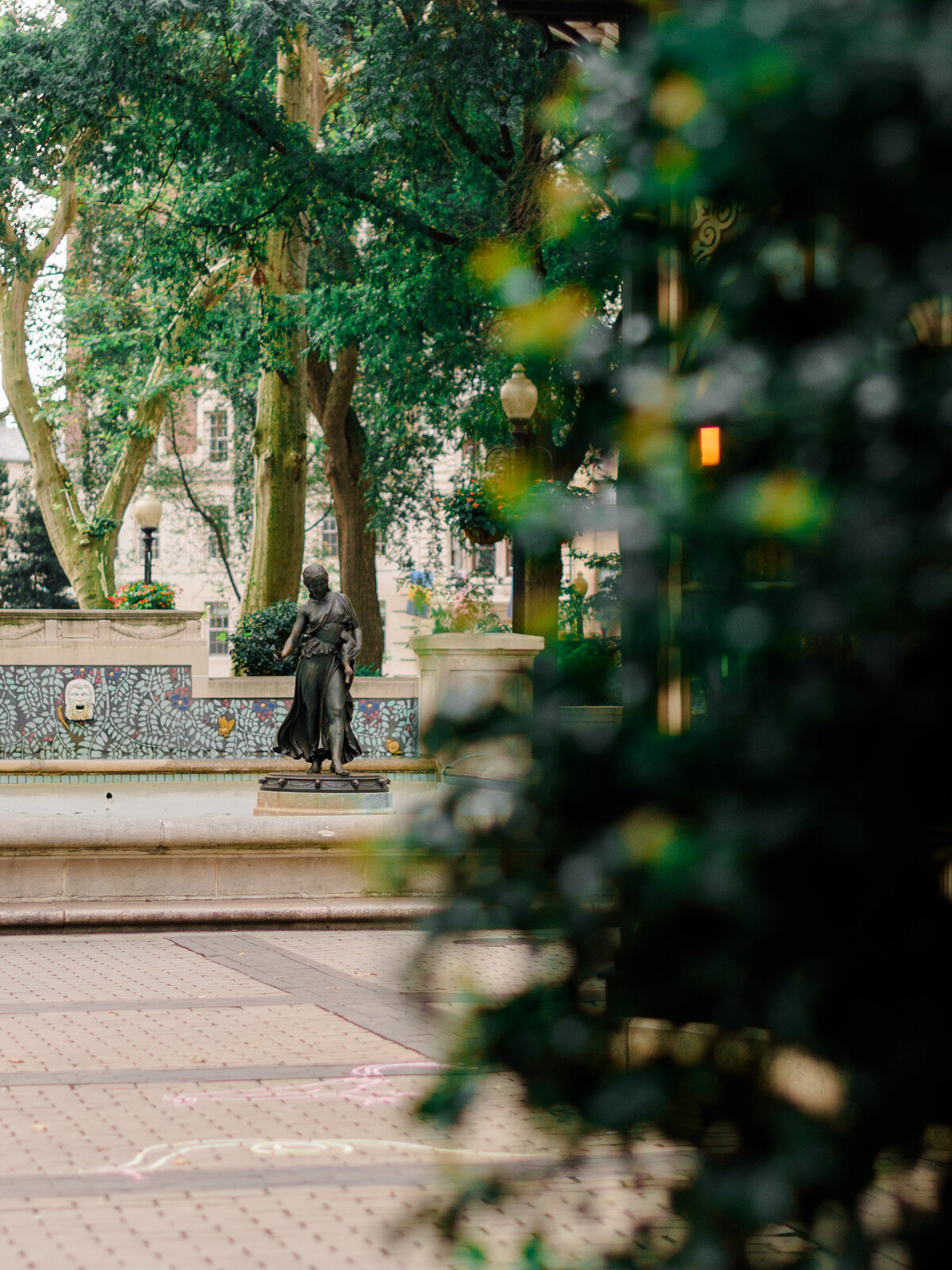 A bronze statue of a woman stands gracefully at the edge of a tiled fountain in Rittenhouse Square, partially framed by soft greenery and dappled light. The surrounding mosaic wall and elegant architecture hint at the park's rich history, captured by Elizabeth Kane Photography, a Luxury Philadelphia Wedding Photographer.