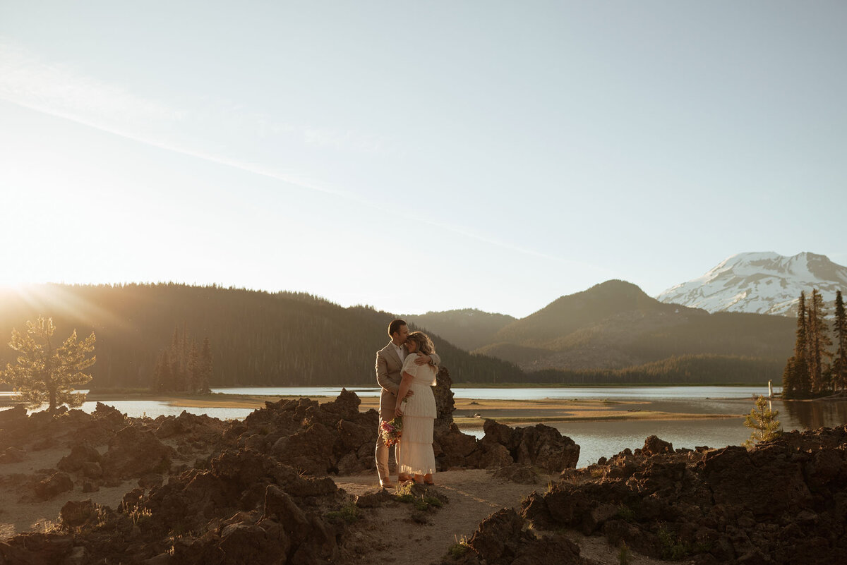 groom hugs and kisses his bride while standing next to Sparks Lake under a clear, blue sky