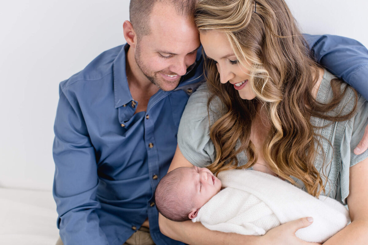 A smiling couple sits closely together, lovingly holding a newborn baby wrapped in a soft white blanket. The man is wearing a blue shirt, and the woman has long, wavy hair and is dressed in light-colored clothing.