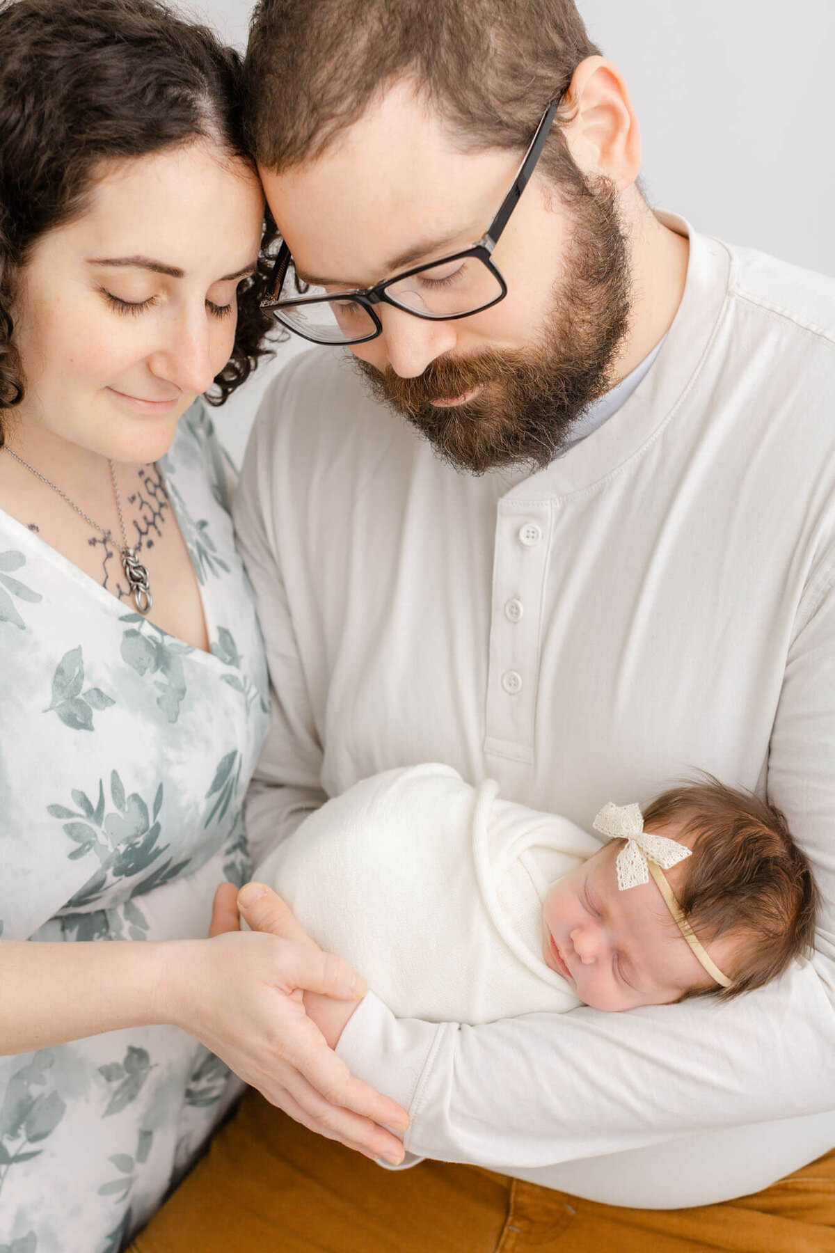 Dad is holding new baby girl in arms and Mom is standing next him. Mom and Dad's foreheads are touching. Baby is wrapped in white and sleeping peacefully. Mom is wearing a green floral dress and dad is wearing brown pants and a white henley.