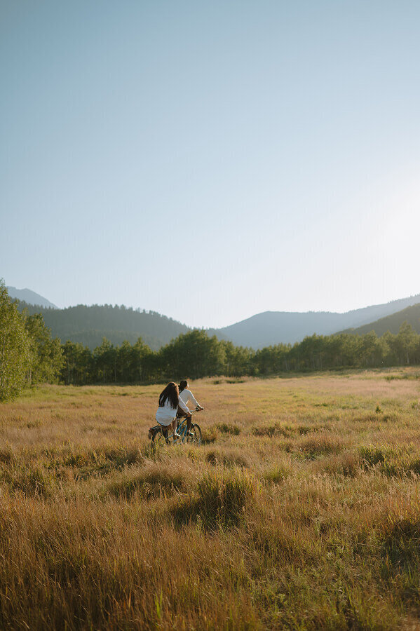 A woman and her girlfriend ride a bicycle through a field of tall grass during sunset in Swan Valley ID.