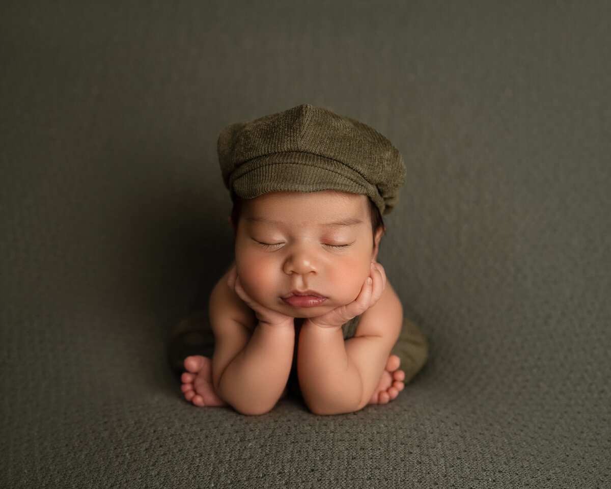 Baby boy in classic froggy pose wearing green cap on seamless olive background | Studio newborn photography in Cedar Park and Round Rock